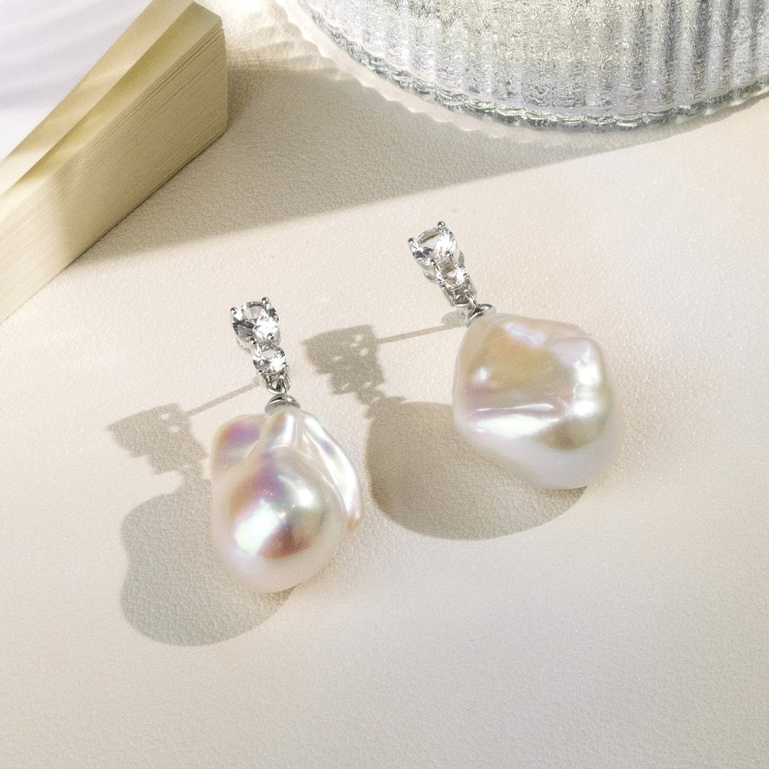Baroque iridescent freshwater pearl double CZ drop earrings on cream surface flatlay with glass prop