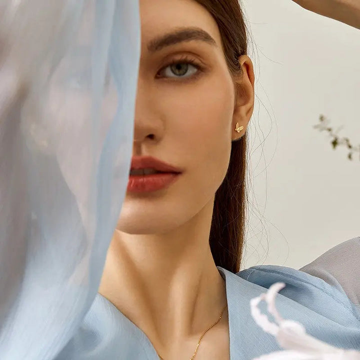 Model wearing gold vermeil butterfly stud earring in 925 sterling silver, artistic portrait with blue sheer fabric